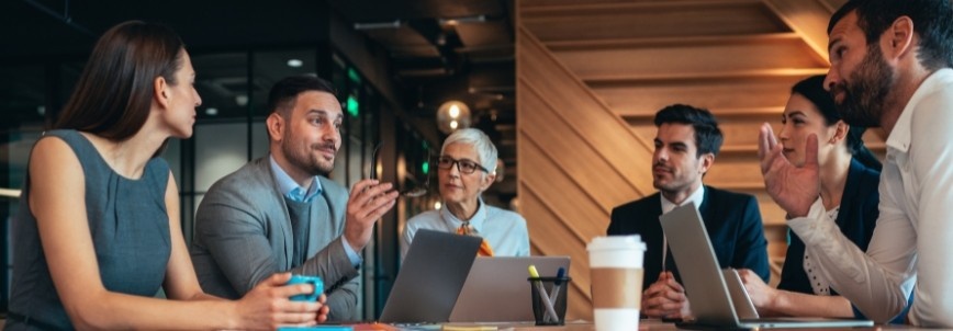 Six business professionals sit around a table in a modern office, engaged in a focused discussion about what is development approach. Laptops, papers, and a coffee cup are on the table as they share ideas and strategies.