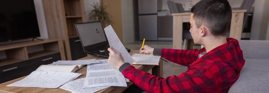 A person in a red plaid shirt sits at a table covered with papers, notebooks, and a laptop, using a pencil to study for the PMP Simulator in a cozy living room setting.