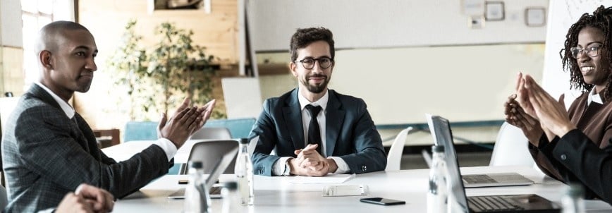Four professionals in business attire sit around a conference table. As three applaud, the man in the center—perhaps pondering why get the CAPM certification—smiles with hands clasped. Laptops and papers are spread out before them.