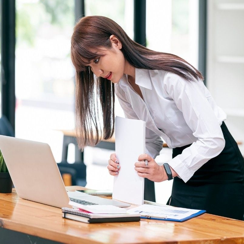 A woman in a white shirt and black skirt leans over a desk, holding a document while looking at a laptop—engaged in CAPM Exam Preparation. Papers, a notebook, and a pen are on the wooden desk in a bright office setting.