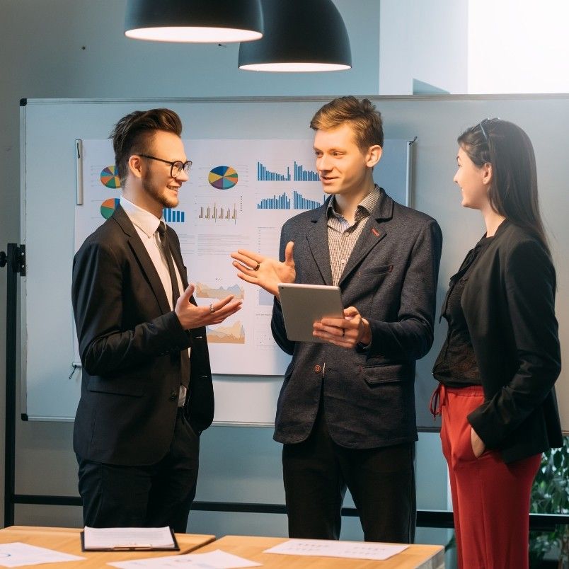 Three professionally dressed people stand in front of a whiteboard with charts, engaged in discussion about types of Agile projects. One holds a tablet, another gestures while speaking, and the third person listens and smiles.
