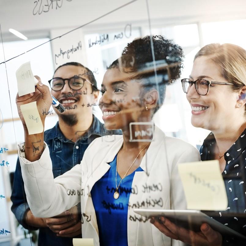 Three people smile and collaborate, writing on a glass wall with sticky notes and diagrams while discussing Types of Agile Projects. One person writes with a marker as the others watch, holding notes and a tablet in a bright office setting.