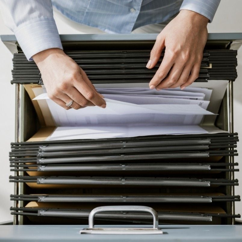 A person’s hands are sorting through hanging file folders in an open filing cabinet drawer, organizing or searching for documents such as a Project Charter.