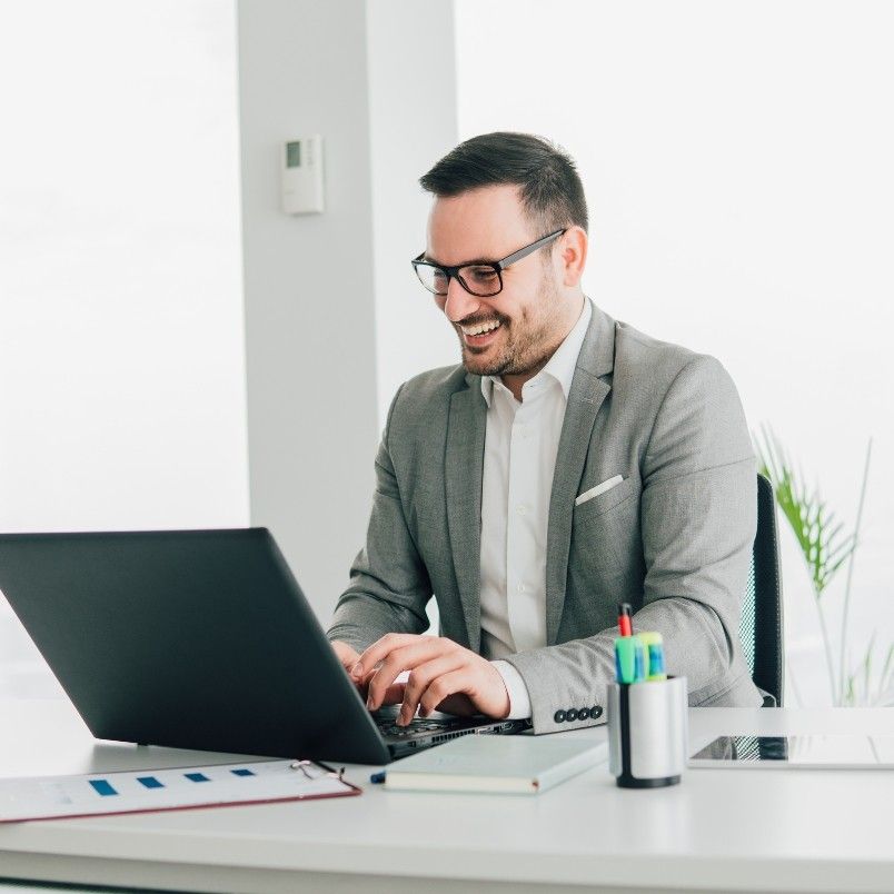 A smiling man in a gray suit and glasses works on a laptop at a modern, bright office desk, researching why get the CAPM certification. Office supplies, papers, and a tablet are on the white desk.