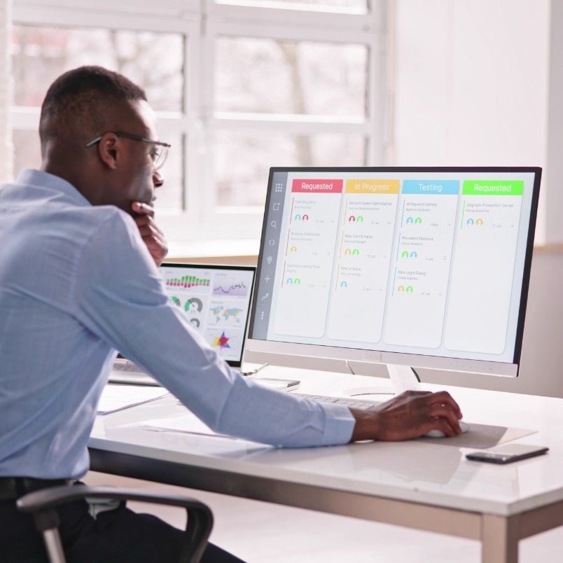 A person sits at a desk, studying for their CAPM Certification while examining a large computer monitor displaying a project management dashboard with columns labeled Requested, In-Progress, Testing, and Requested. Daylight streams in through a window in the background.