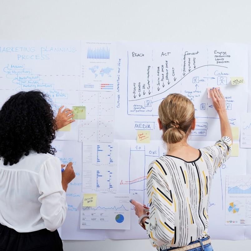 Two women stand facing a wall covered with charts, graphs, and notes, discussing types of Agile projects as they collaborate on strategy and analysis, adding sticky notes to a marketing planning board.