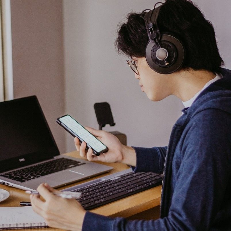 A person wearing headphones sits at a desk with a laptop, focused on CAPM Exam Preparation. They hold a pen in one hand and look at their phone, surrounded by papers and a cup.