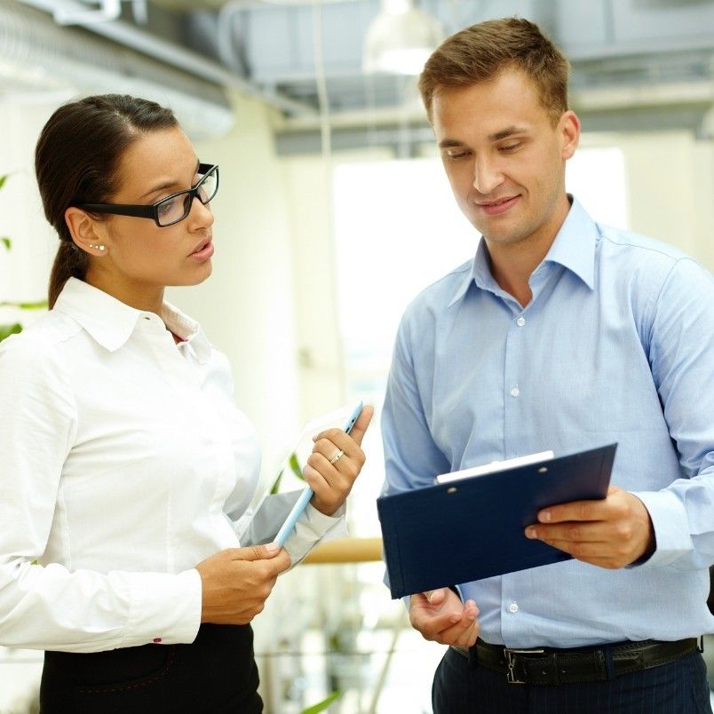 Two business professionals stand indoors, discussing the pros and cons of onsite PMP testing. The woman, wearing glasses and a white shirt, holds a tablet, while the man in a light blue shirt smiles at a clipboard.