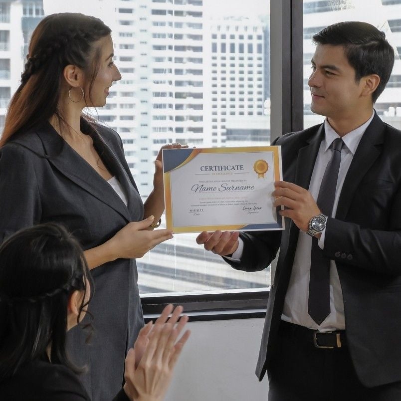 A man in a suit hands a certificate to a woman in business attire, recognizing her CAPM Exam Preparation success, while another woman applauds. They are standing in an office with large windows overlooking city buildings.
