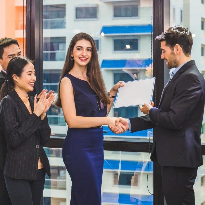 A woman in a blue dress receives a certificate for CAPM Exam Preparation and a handshake from a man in a suit, while two colleagues stand nearby, smiling and clapping, in an office setting with large windows.
