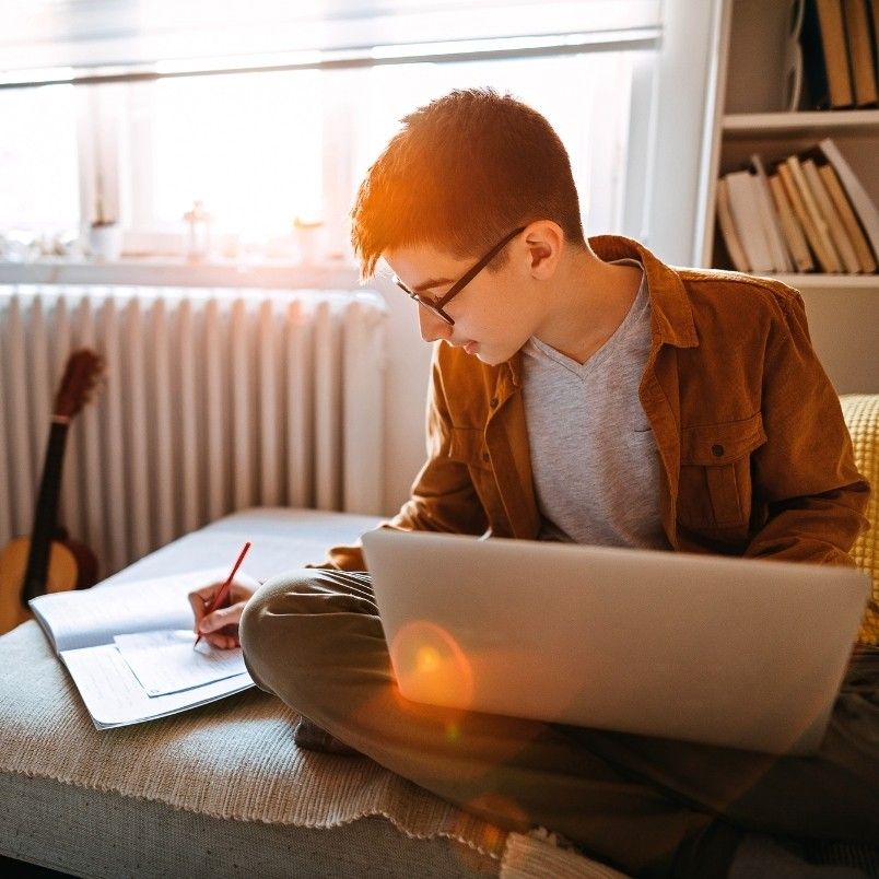 A boy wearing glasses sits cross-legged on a couch, using a laptop and writing in a notebook—perhaps reflecting on the ups and downs of online PMP® exams. Sunlight streams through the window, with a guitar and bookshelves in the background.