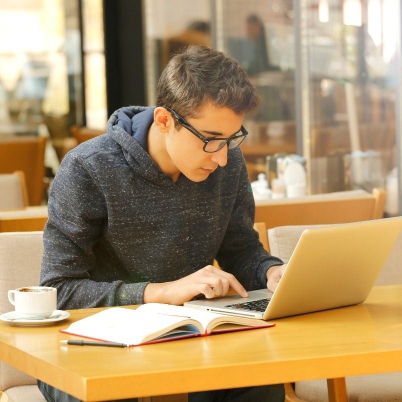 A young person wearing glasses and a hoodie is sitting at a cafe table, focused on CAPM Exam Preparation on their laptop. An open notebook and a cup of coffee are on the table beside them.