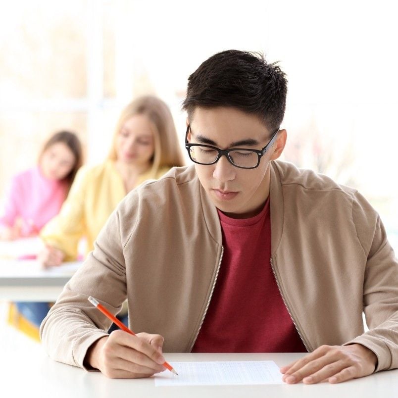 A young man wearing glasses and a beige jacket writes on a sheet of paper at a desk, focused on his CAPM Exam Preparation. Two other students are seated and writing in the background in a bright classroom.