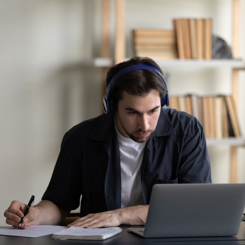 A young man wearing headphones sits at a desk, looking intently at a laptop screen while writing notes on paper—perhaps researching why get the CAPM certification. Bookshelves filled with books are visible in the background.
