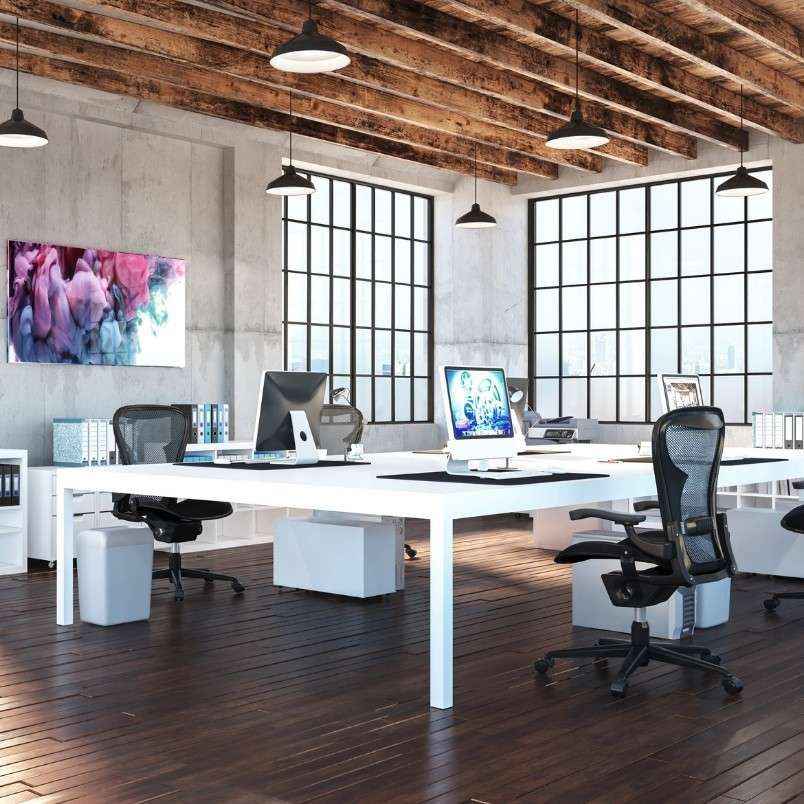 Modern open-plan office with large windows, white desks, black mesh chairs, desktop computers, and colorful abstract art—a bright space ideal for discussing the Pros and Cons of Onsite PMP Testing in a collaborative environment.