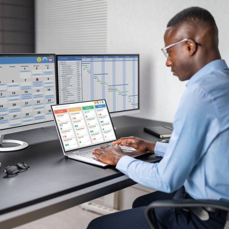 A man in business attire works at a desk with a laptop and two large monitors displaying a calendar, a Gantt chart, and a CAPM Exam Preparation project management dashboard.