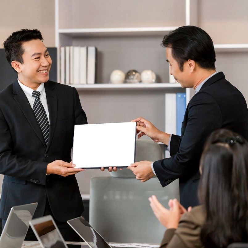 Two men in business suits smile while one hands a blank white certificate—possibly discussing CAPM® Certification Cost—to the other in an office setting, as colleagues seated nearby applaud.
