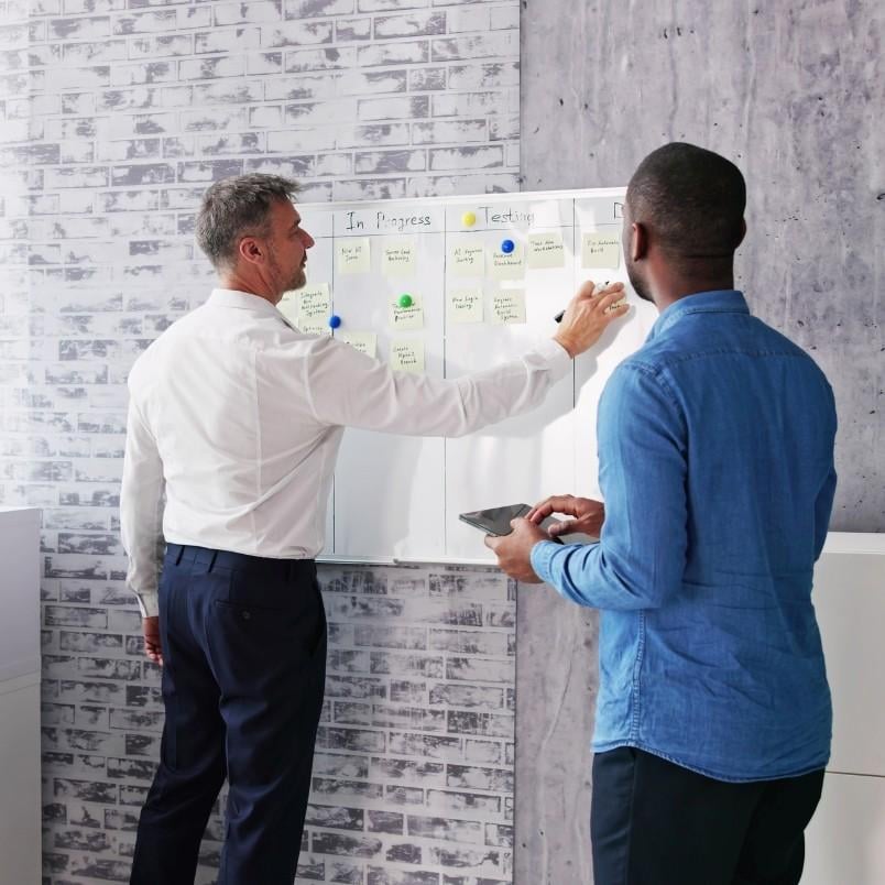 Two men stand in front of a whiteboard with sticky notes organized in columns; one, likely a Project Manager in Agile, writes on the board while the other holds a tablet and observes.