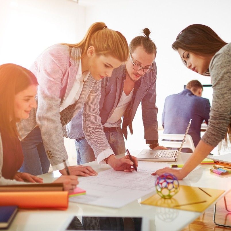 Four young adults collaborate around a table, reviewing documents and taking notes as they discuss Types of Agile Projects. One person uses a laptop, while others lean in to share ideas. Office supplies and a decorative object are on the table.