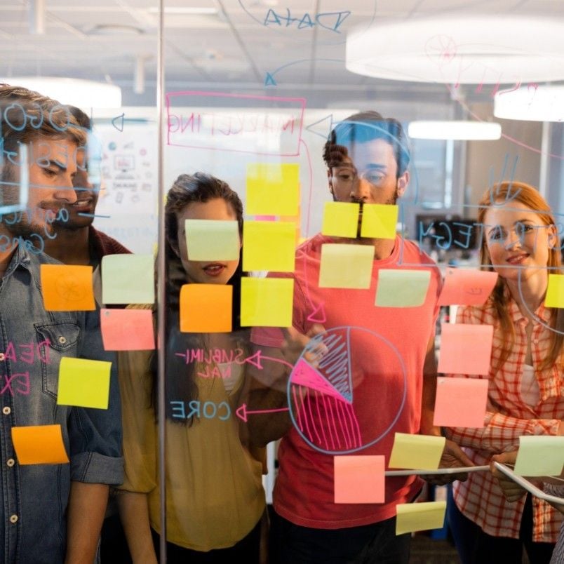 Four people, including a Project Manager in Agile, stand behind a glass wall covered with colorful sticky notes and diagrams, collaborating and discussing ideas in a modern office setting.