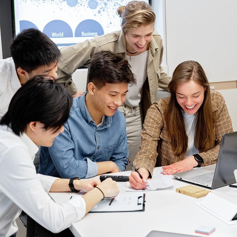 Five young adults gather around a table, smiling and collaborating on paperwork and a laptop, suggesting a team meeting or group study session discussing Types of Agile Projects in a modern, bright office or classroom.