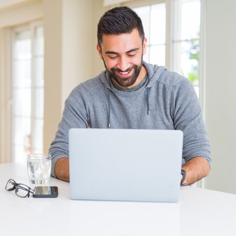 A smiling man with a beard sits at a white table using a laptop, perhaps navigating the ups and downs of online PMP® exams. A glass of water, eyeglasses, and smartphone rest beside him, with bright windows in the background.
