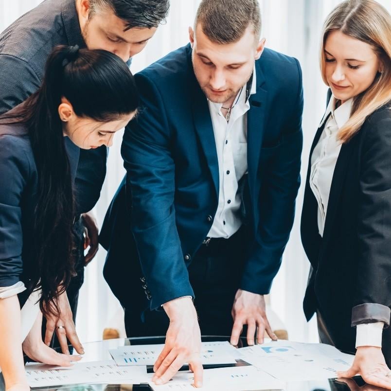 Four business professionals in formal attire stand around a table, closely examining charts and graphs related to CAPM Exam Preparation, indicating a collaborative discussion or meeting.
