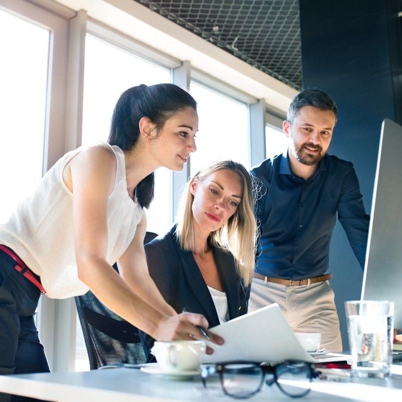 Three people in business attire collaborate at a modern office desk, focused on CAPM Exam Preparation. One woman points at a document, while another woman and a man review it together on the computer screen.
