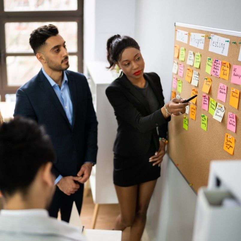 Three professionals in business attire, including a Project Manager in Agile, discuss colorful sticky notes on a corkboard during a meeting. One woman points at a note while two colleagues listen attentively.