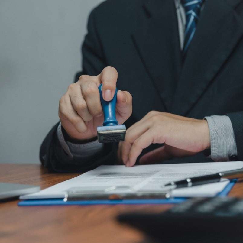 A person in a suit stamps a document on a clipboard at a desk, with CAPM® Certification Requirements materials, a laptop, and calculator visible in the foreground.