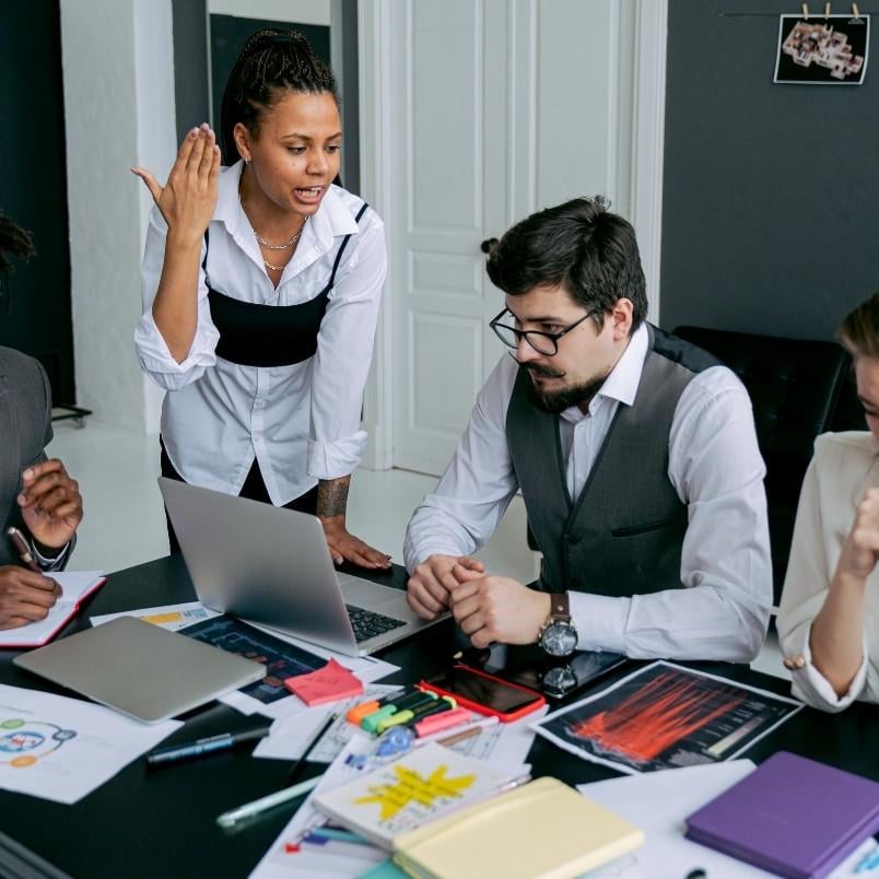 A group of four professionals sits at a cluttered table with laptops and papers during a rosemet CAPM training session. One woman stands and gestures while talking, as the others listen and take notes in a modern office setting.