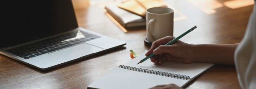 A person writes in a spiral notebook with a pencil at a wooden desk, next to an open laptop displaying PMP® Practice Exams, with a mug and some notebooks in a softly lit setting.