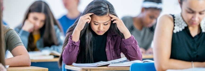 A group of students sits at desks in a classroom, focused on their work. The student in the center looks stressed, possibly feeling PMP Exam Stress, as she holds her head with both hands while reading a notebook.
