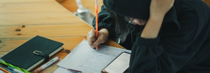 A person wearing a black hoodie sits at a wooden table, writing notes on paper with a pen—preparing strategies on how to pass the PMP exam. An open notebook, a smartphone, and several highlighters are also on the table.