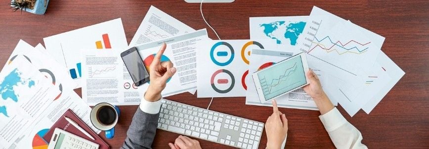 Two people discuss data charts and graphs—essential decision making tools—on paper, a tablet, and a smartphone spread across a desk with a computer keyboard, documents, coffee, and office supplies.