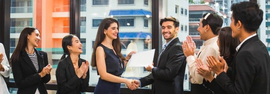 A group of professionally dressed people in an office applauds as a smiling man shakes hands with a woman, congratulating her on meeting CAPM® Certification Requirements and awarding her a certificate. They stand before large windows overlooking city buildings.