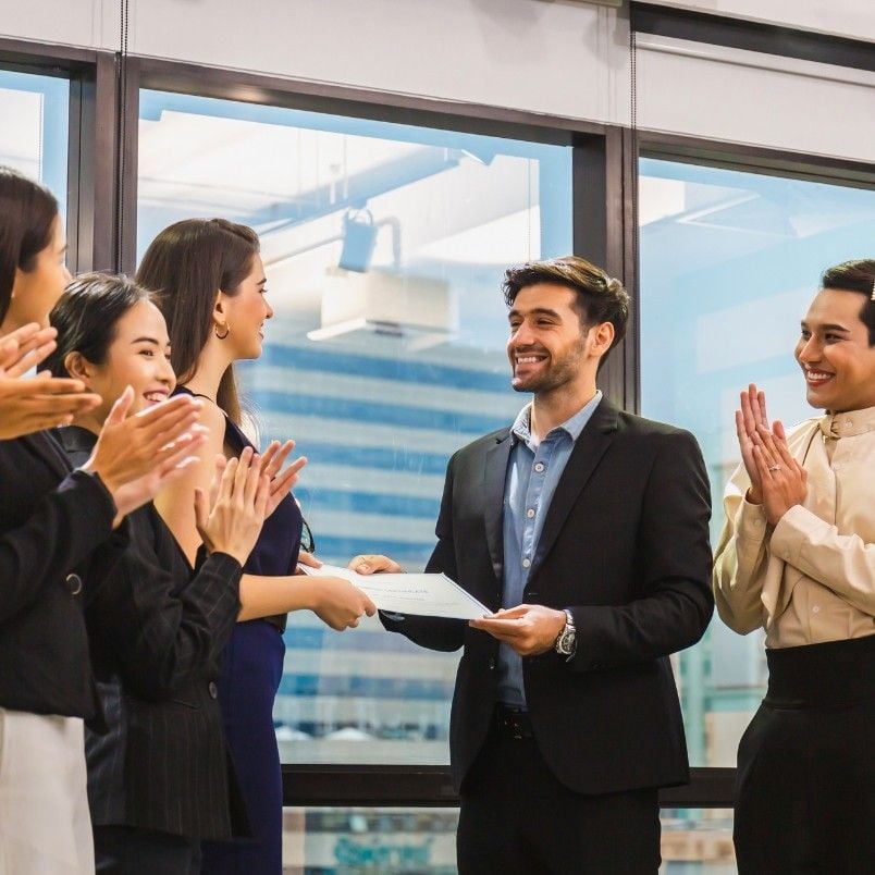 A man in a suit receives a certificate recognizing CAPM® Certification Requirements from a woman, while colleagues stand around them clapping and smiling in an office with large windows.