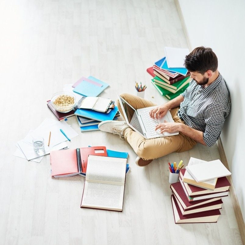 A man sits on the floor with a laptop, surrounded by stacks of books, notebooks, and colored pencils, researching CAPM® Certification Requirements as he studies or works, with a bowl of cereal nearby.