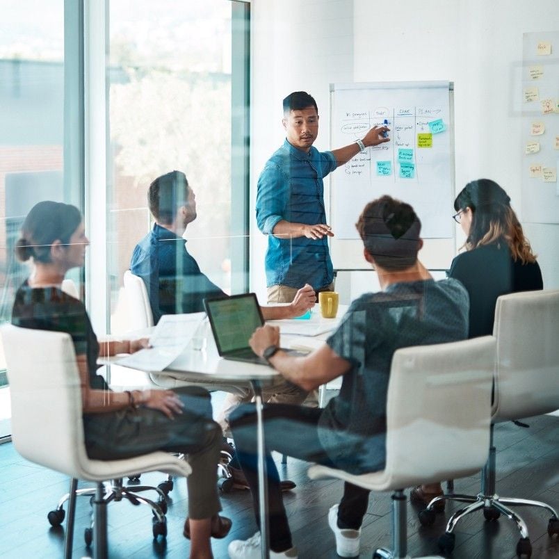 A man stands by a whiteboard with colorful sticky notes, presenting rosemet capm training to four colleagues seated around a conference table with laptops and papers in a bright, modern office.
