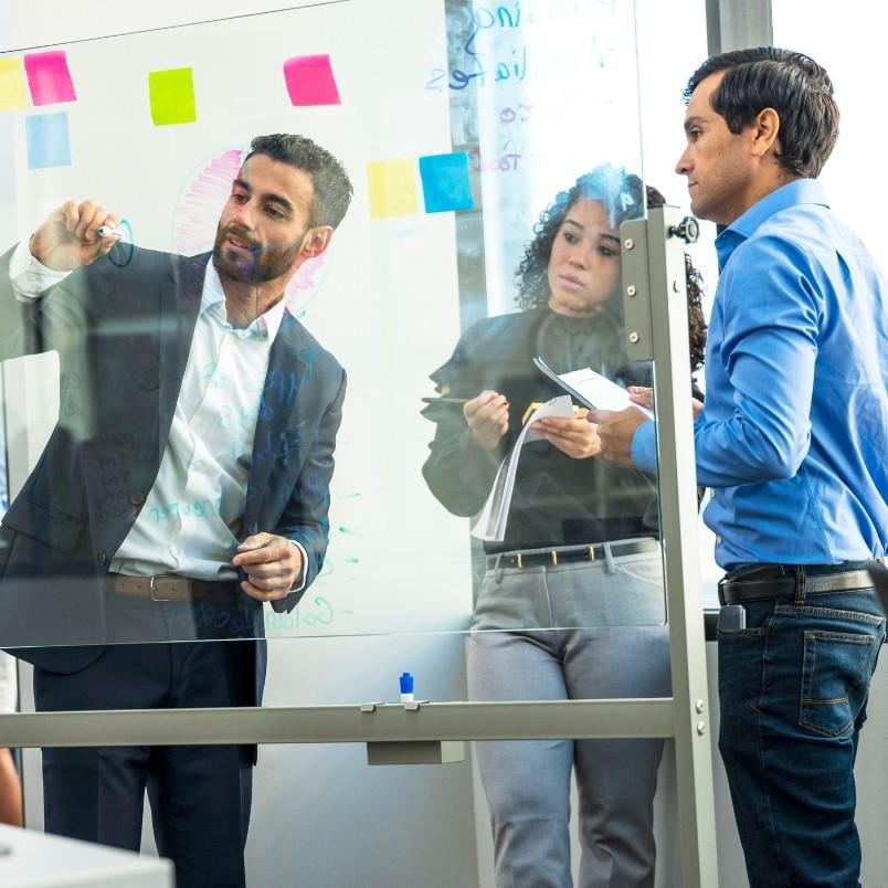Three people collaborate in an office; during a rosemet capm training session, one man writes on a transparent board while a woman with notes and another man watch and discuss. Colorful sticky notes are visible on the board.