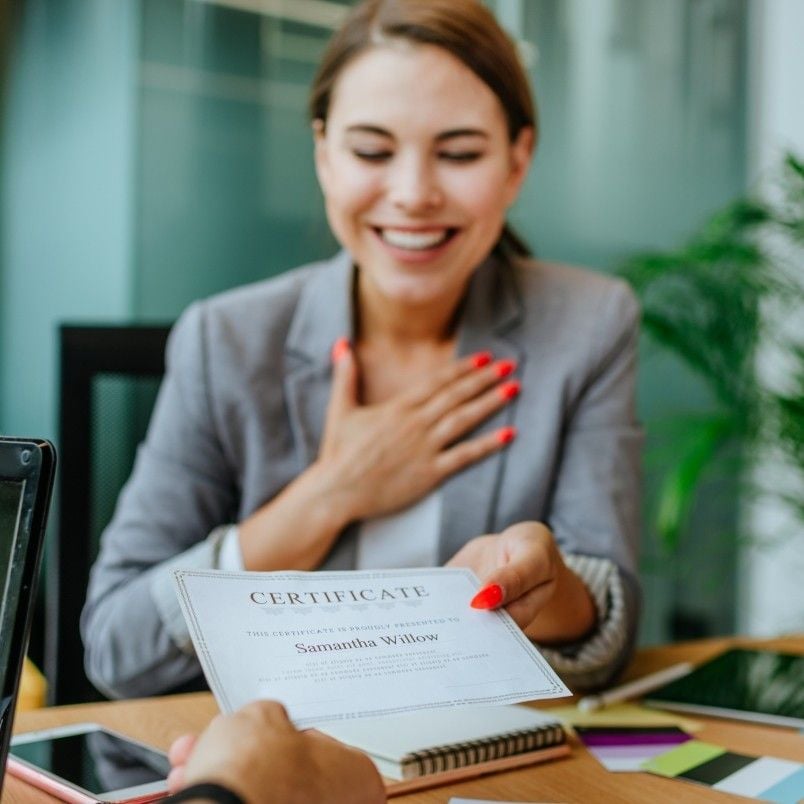 A smiling woman with red nails, holding a certificate and placing her other hand on her chest, sits at a desk during what appears to be an award moment, likely celebrating meeting her CAPM® Certification Requirements.