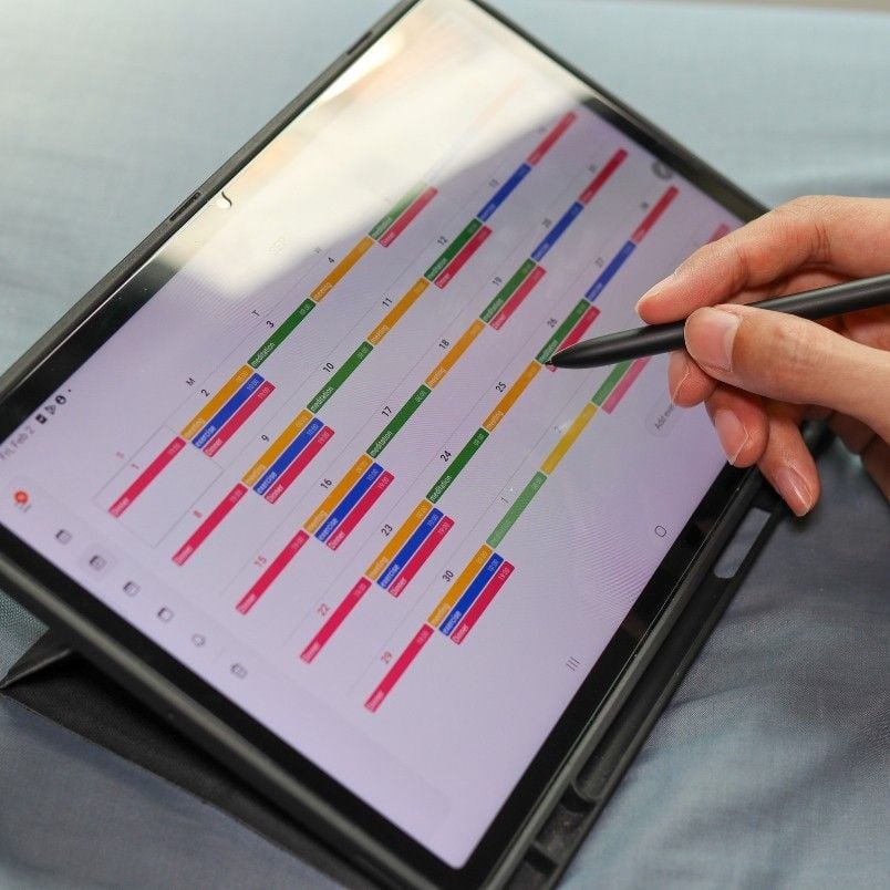 A person uses a stylus to interact with a digital calendar displayed on a tablet, color-coding their monthly schedule to help manage PMP Exam stress.