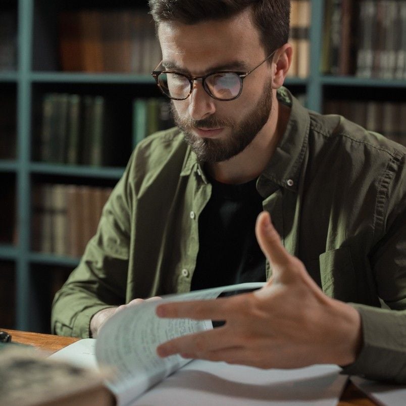 A man wearing glasses and a green shirt sits at a table in a library, reading and flipping through pages of a notebook as he prepares for the PMP Exam, stress evident against the backdrop of bookshelves.