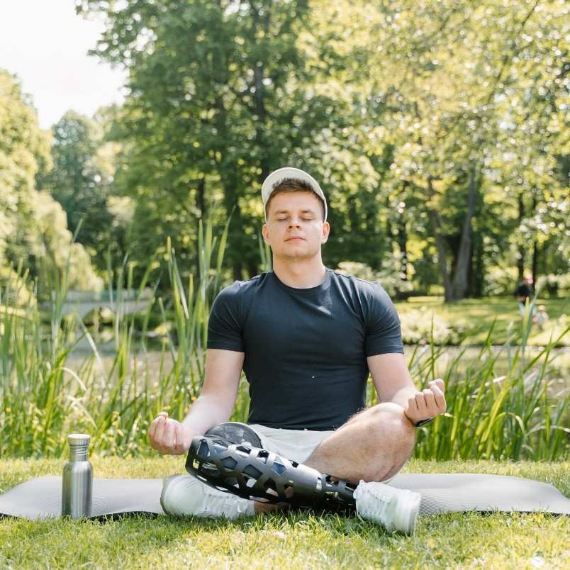 A man with a prosthetic leg sits cross-legged on a mat outdoors, meditating with eyes closed. Amid green grass and sunlight, he finds calm in the park, managing PMP Exam Stress. A water bottle rests beside him.