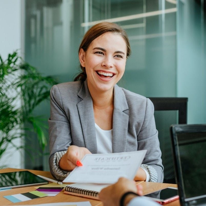 A smiling woman in a gray blazer sits at a desk, handing a resume to someone across the table during a job interview, offering encouragement for overcoming PMP Exam Stress in a modern office setting.