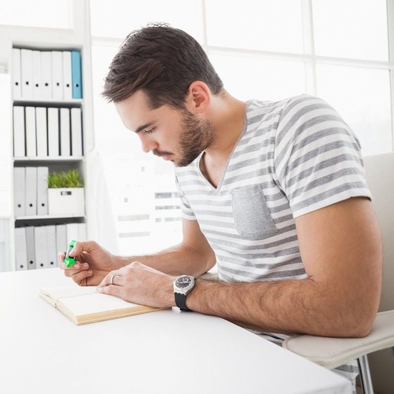 A man wearing a striped t-shirt sits at a desk, writing notes about CAPM® Certification Requirements with a green pen. He appears focused, while shelves with binders and a small plant are visible in the background.