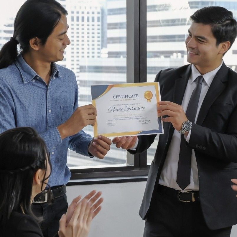 Two men stand indoors by a window, one in a blue shirt and one in a suit, smiling as they hold a certificate recognizing CAPM® Certification Requirements while two seated people clap. Urban buildings are visible outside.