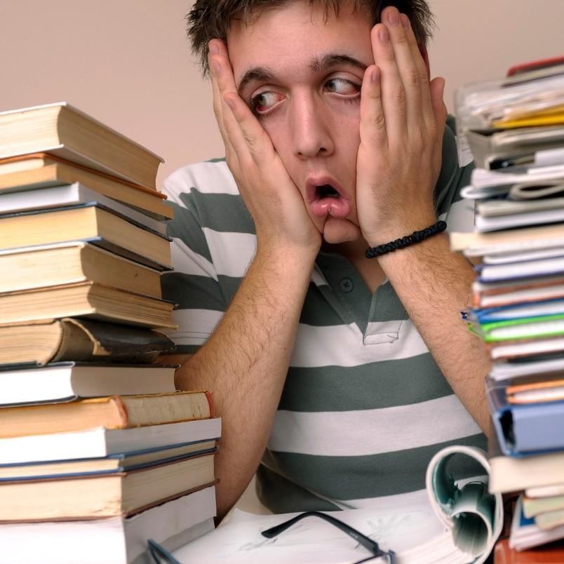 A young person in a striped shirt looks overwhelmed, holding their face with both hands, surrounded by large stacks of books and papers on a desk—clearly feeling the weight of PMP Exam Stress.