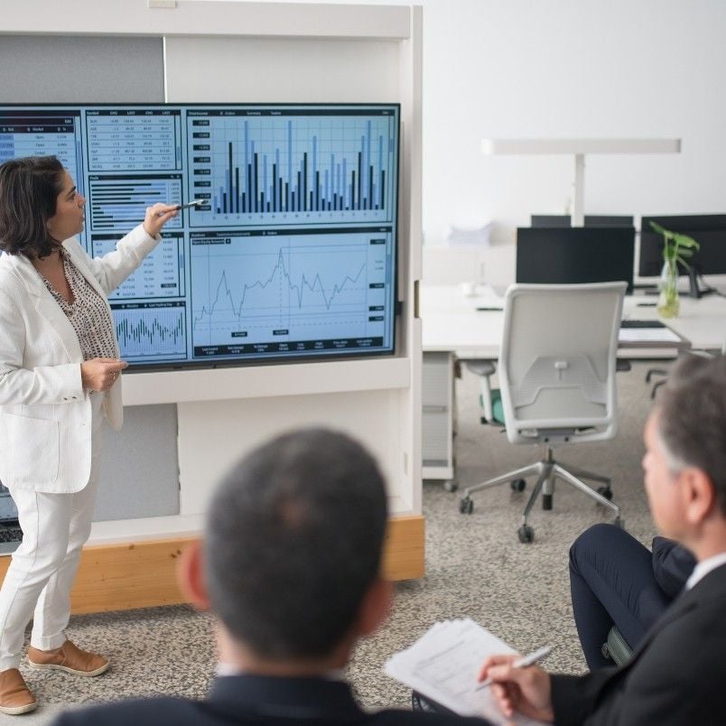 A woman in a white suit is presenting graphs and charts on large screens, showcasing alternative analysis to two seated colleagues in a modern office setting.