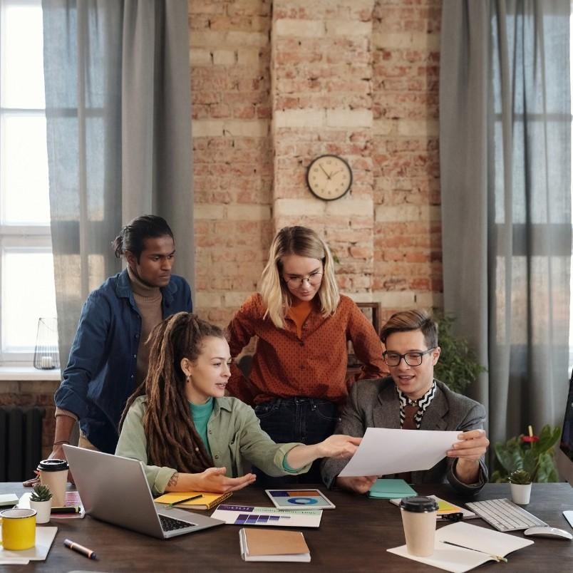 Four people gather around a table with laptops, papers, and coffee cups, collaborating on a document in a modern office—a great agile project management example set against brick walls and large windows.